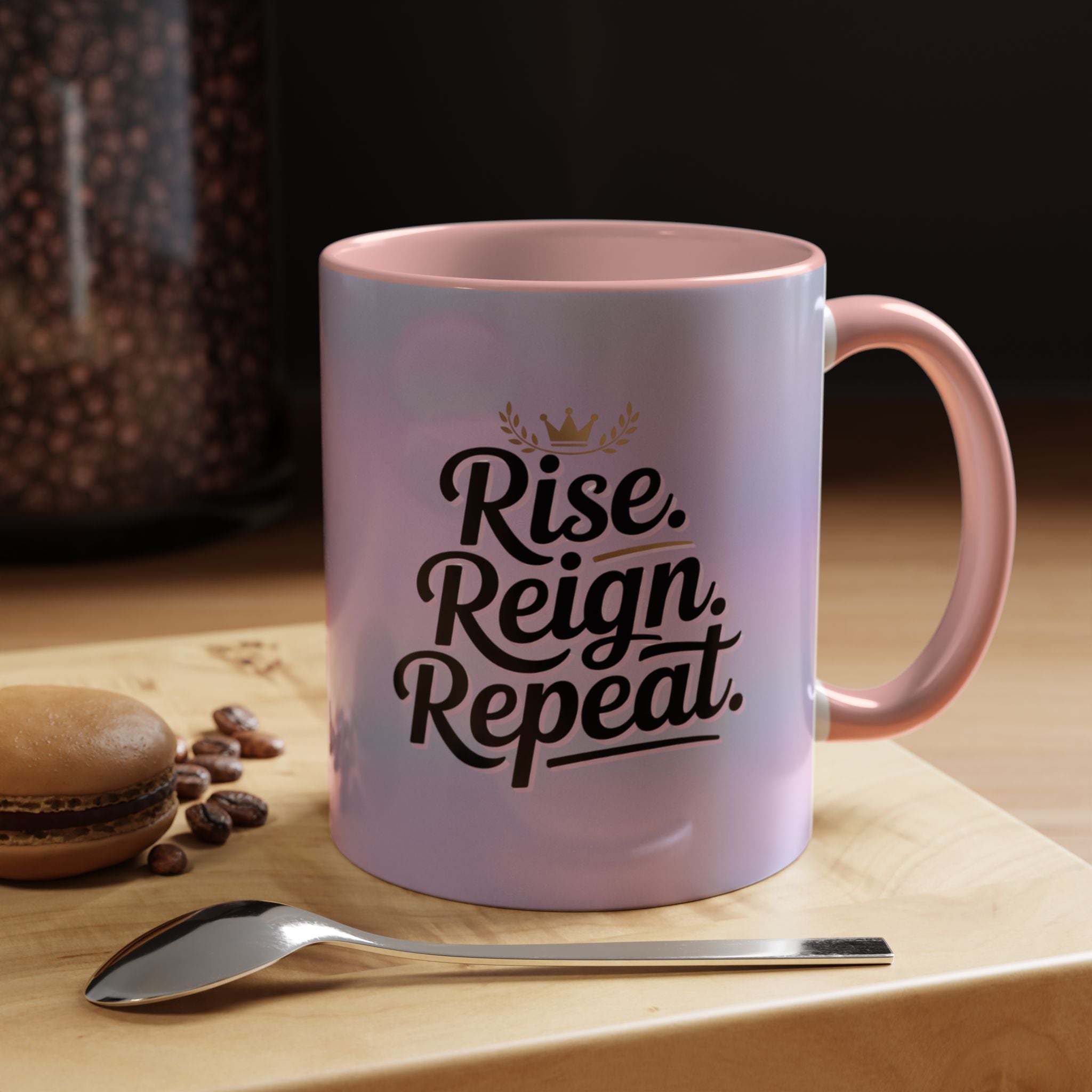Pink mug with 'Rise. Reign. Repeat.' text on a wooden surface with coffee beans and a spoon.
